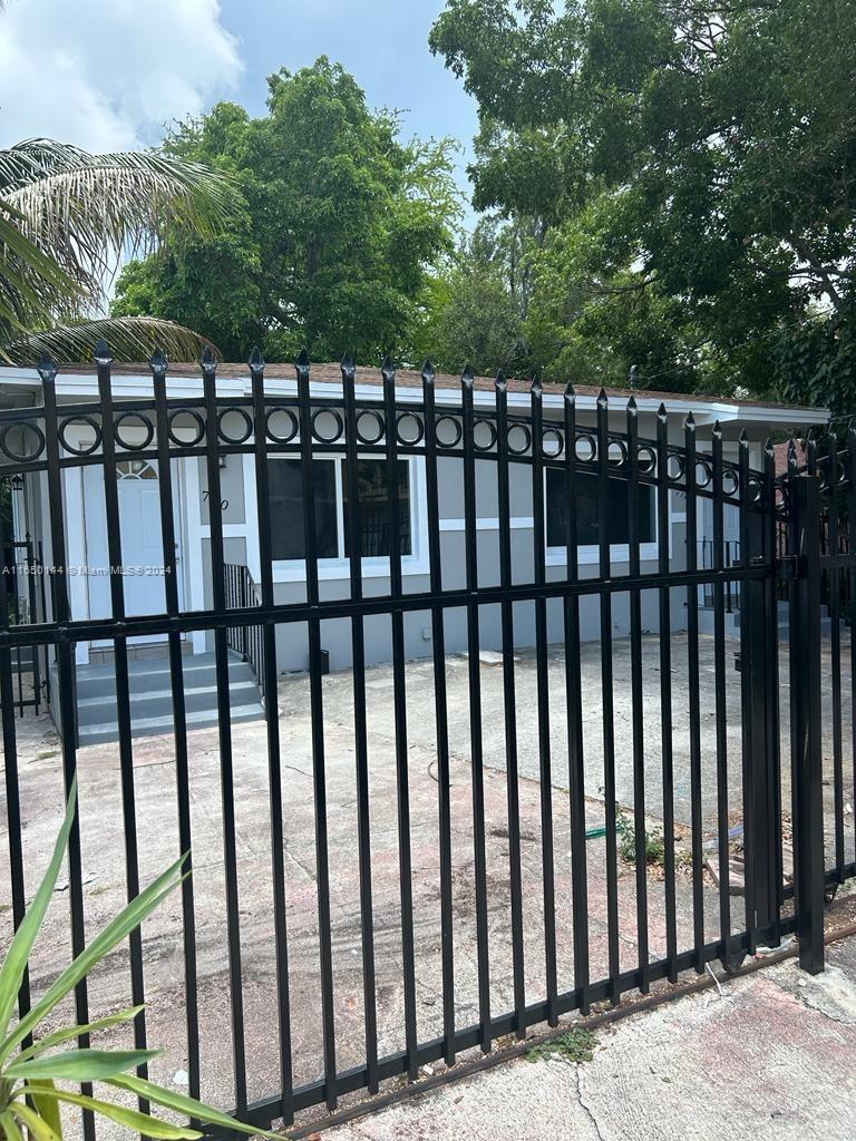 A single-story house with gray siding is viewed behind a black metal security fence — 7710 NE 1st Ct, Miami FL 33138, MIAMI, FL