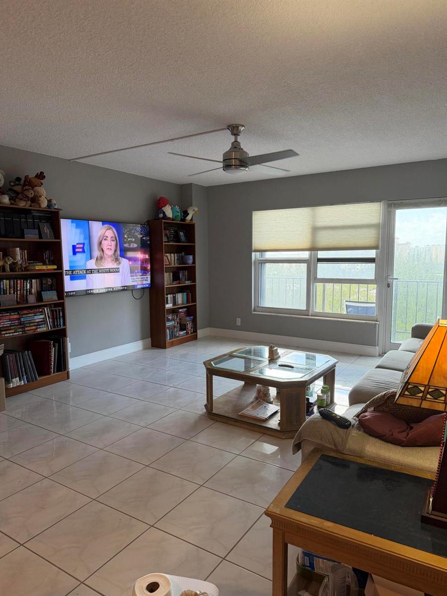 A living room features tile flooring, a ceiling fan, bookshelves, a television, and a balcony door — 405 N Ocean Boulevard # 809, Pompano Beach FL 33062, POMPANO BEACH, FL