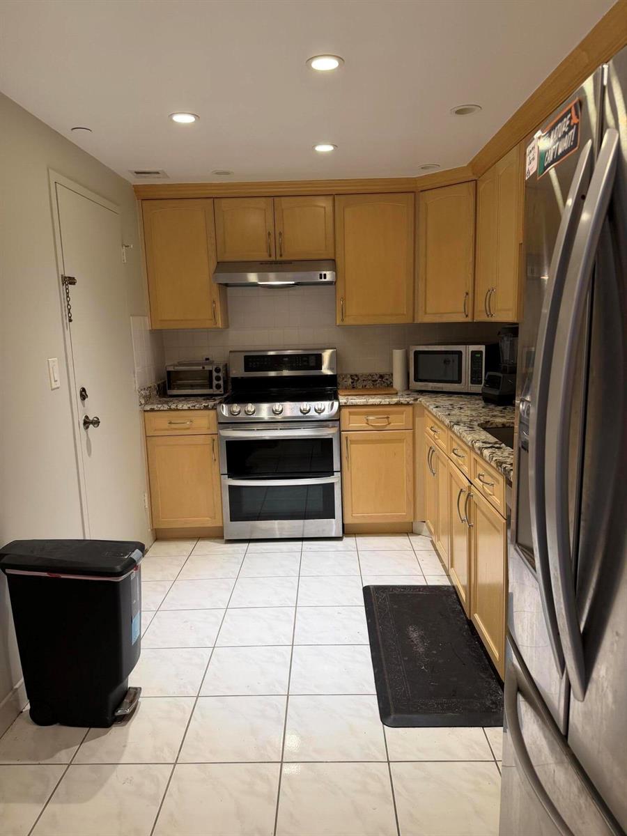 A kitchen features light wood cabinets, stainless steel appliances, granite countertops, and white tile flooring — 405 N Ocean Boulevard # 809, Pompano Beach FL 33062, POMPANO BEACH, FL