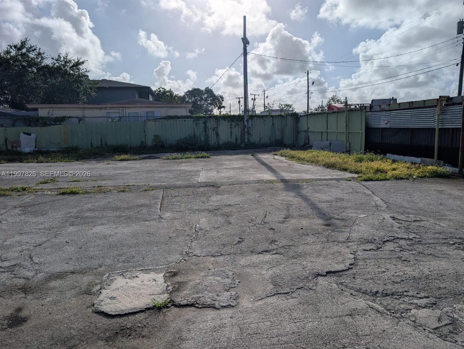 A paved, cracked concrete lot is enclosed by a green metal fence under a cloudy sky — 3175 NW 75TH STREET, Miami FL 33147, MIAMI, FL