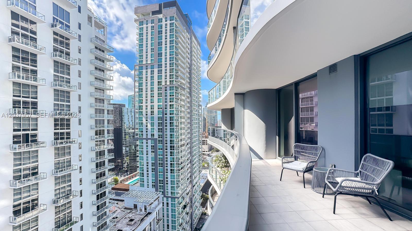 A curved balcony features two gray chairs and a small table overlooking high-rise buildings — 1000 Brickell Plz # 2603, Miami FL 33131, MIAMI, FL
