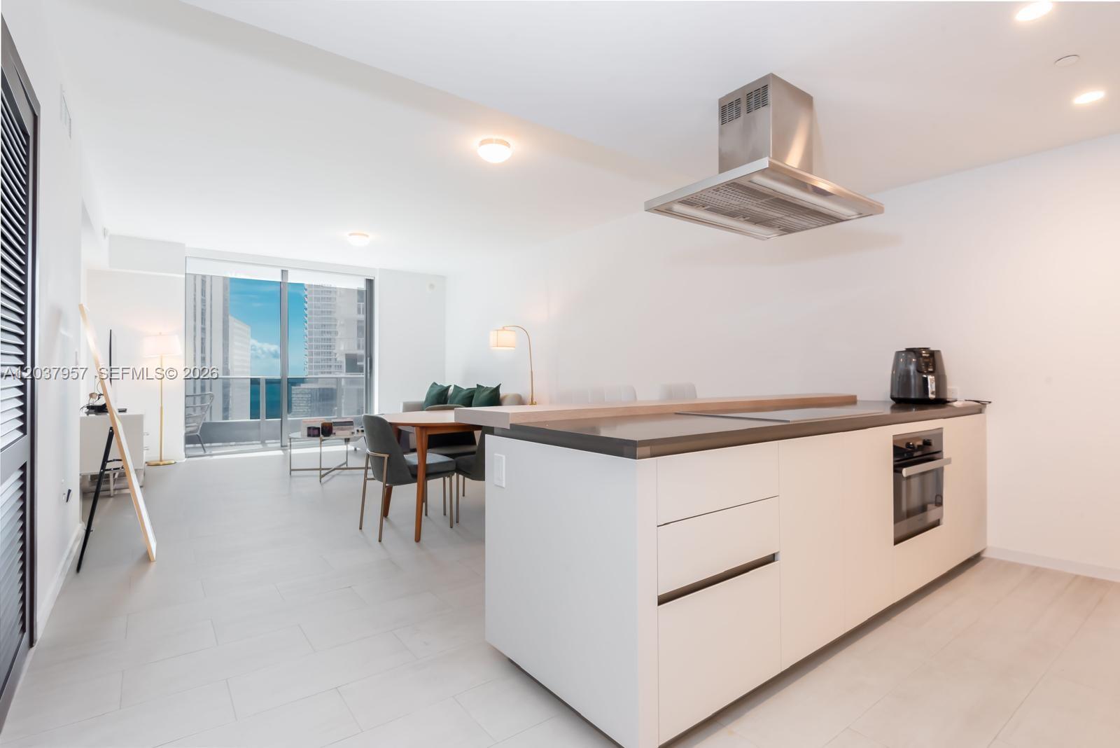 A modern kitchen with a white island and stainless steel hood overlooks a living area — 1000 Brickell Plz # 2603, Miami FL 33131, MIAMI, FL
