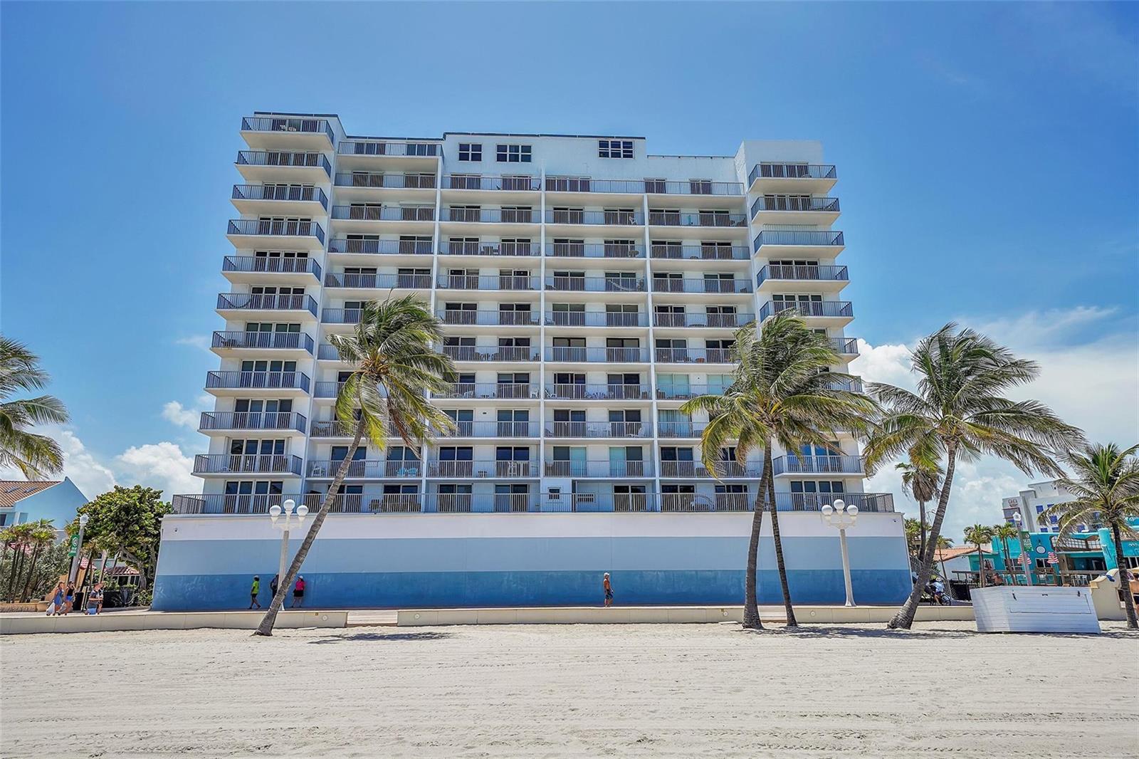 A multi-story beachfront condominium building stands behind a sandy beach with several palm trees — 400 N Surf Road # 703, Hollywood FL 33019, HOLLYWOOD, FL