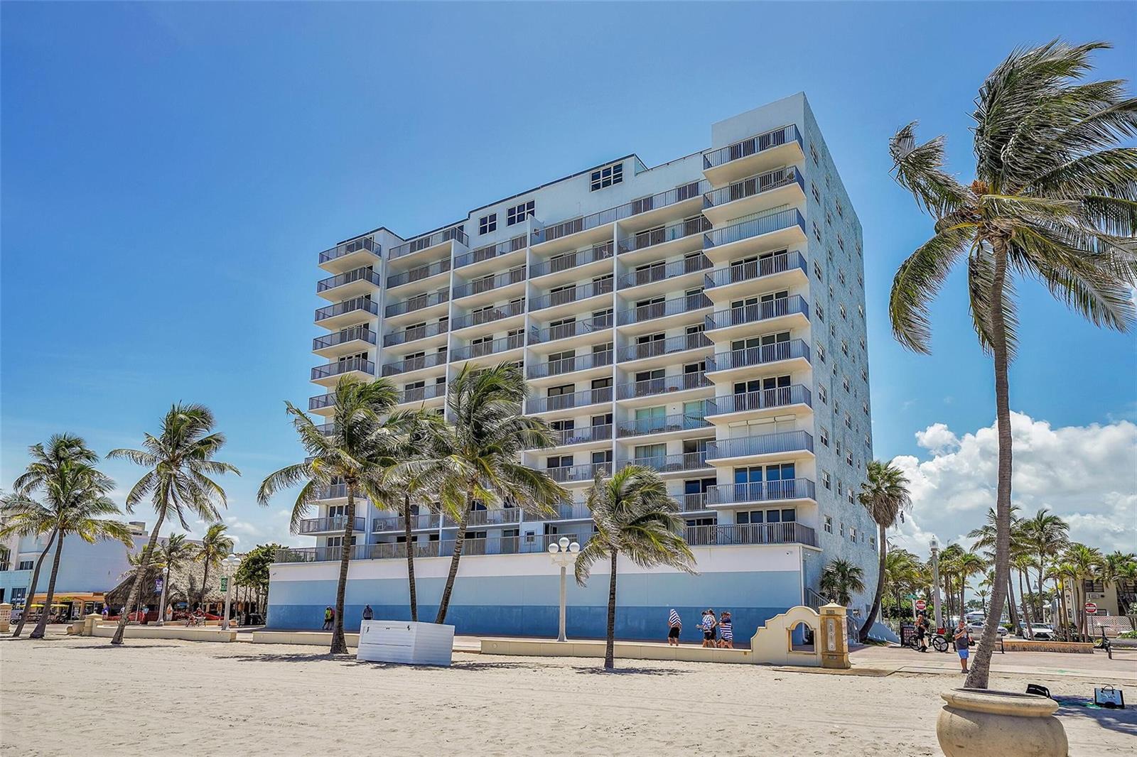 A multi-story beachfront condominium building stands behind a sandy beach with palm trees — 400 N Surf Road # 703, Hollywood FL 33019, HOLLYWOOD, FL