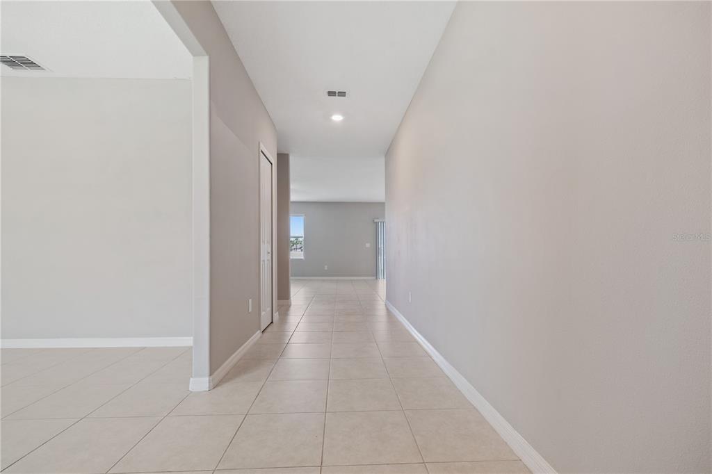 A hallway with light-colored tile flooring leads toward an open living area with windows — 12787 MAPLE BONSAI DRIVE, RIVERVIEW, FL