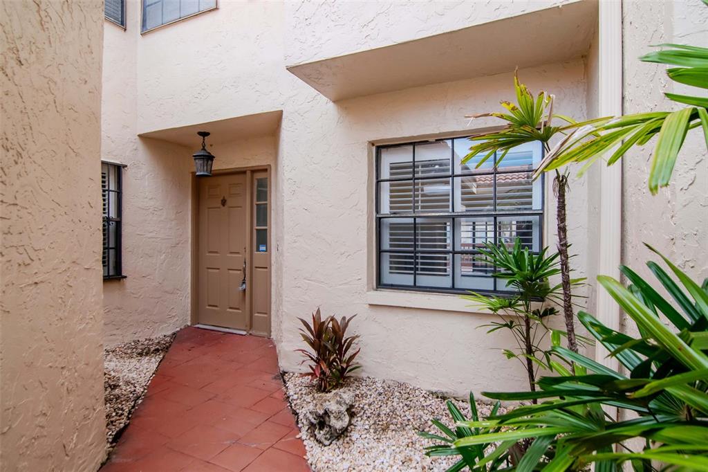 A tan stucco home entrance features a brown front door, tiled walkway, and small garden — 10417 SAINT TROPEZ PLACE UNIT 103, TAMPA, FL