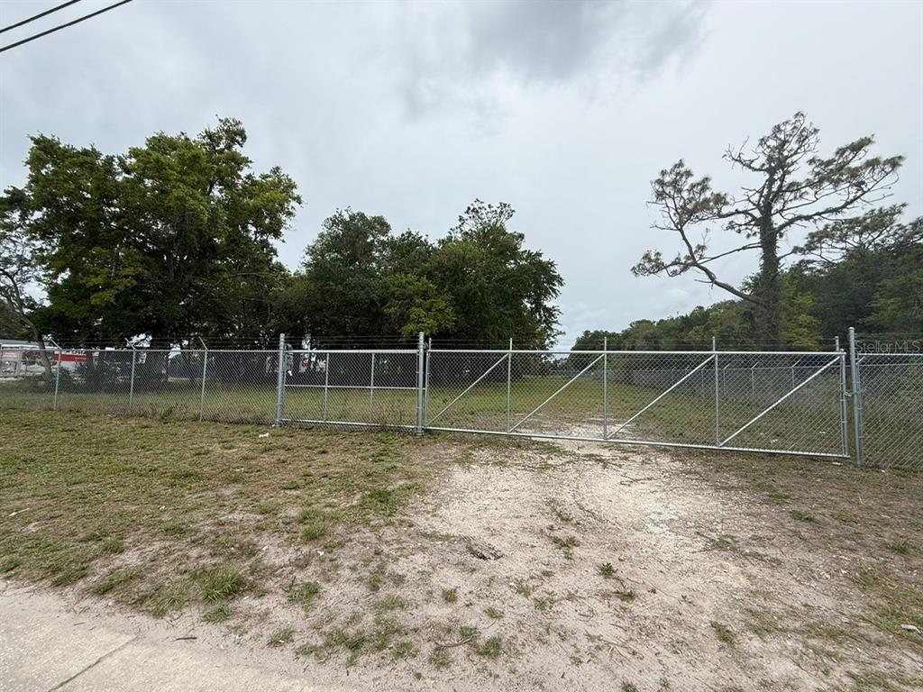 A fenced, gated vacant lot features a dirt driveway and mature trees under a cloudy sky — S GOLDENROD ROAD, ORLANDO, FL