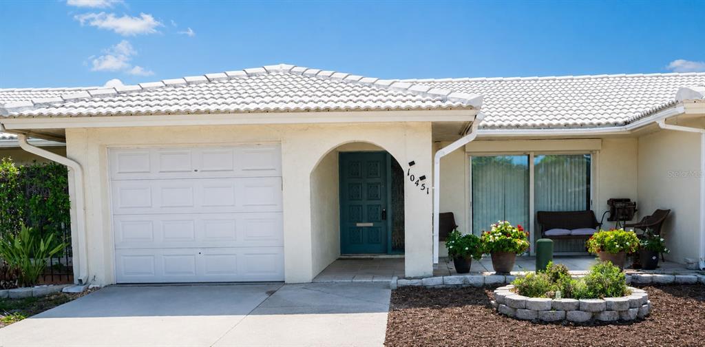 A single-story house features a white garage, a teal front door, and a landscaped yard — 10451 AZALEA PARK DRIVE N UNIT 2, PINELLAS PARK, FL