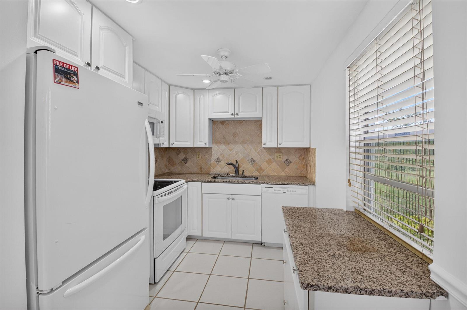 A white kitchen features granite countertops, tile flooring, a ceiling fan, and a window — 200 Normandy E # 200, Delray Beach FL 33484, DELRAY BEACH, FL