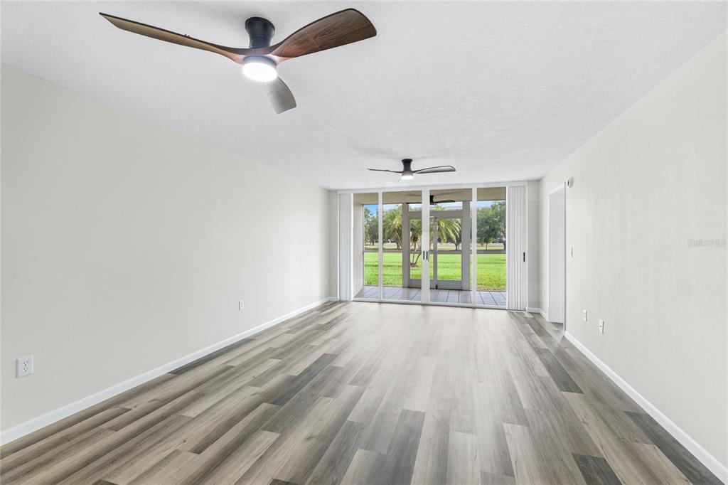 An empty living room features light-colored walls, wood-look flooring, two ceiling fans, and sliding glass doors — 4550 PINEBROOK CIRCLE UNIT 106, BRADENTON, FL