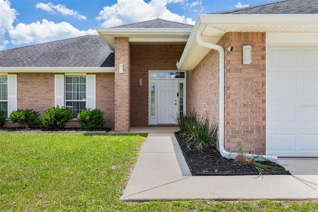 A brick house exterior features a white front door, a concrete walkway, and green landscaping — 34 FOREST GROVE DRIVE, PALM COAST, FL