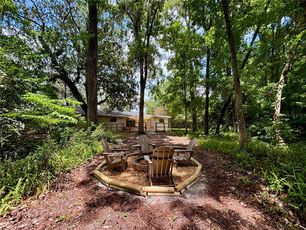 A fire pit area with four Adirondack chairs sits in a wooded backyard behind a house — 4415 NW 33RD COURT, GAINESVILLE, FL