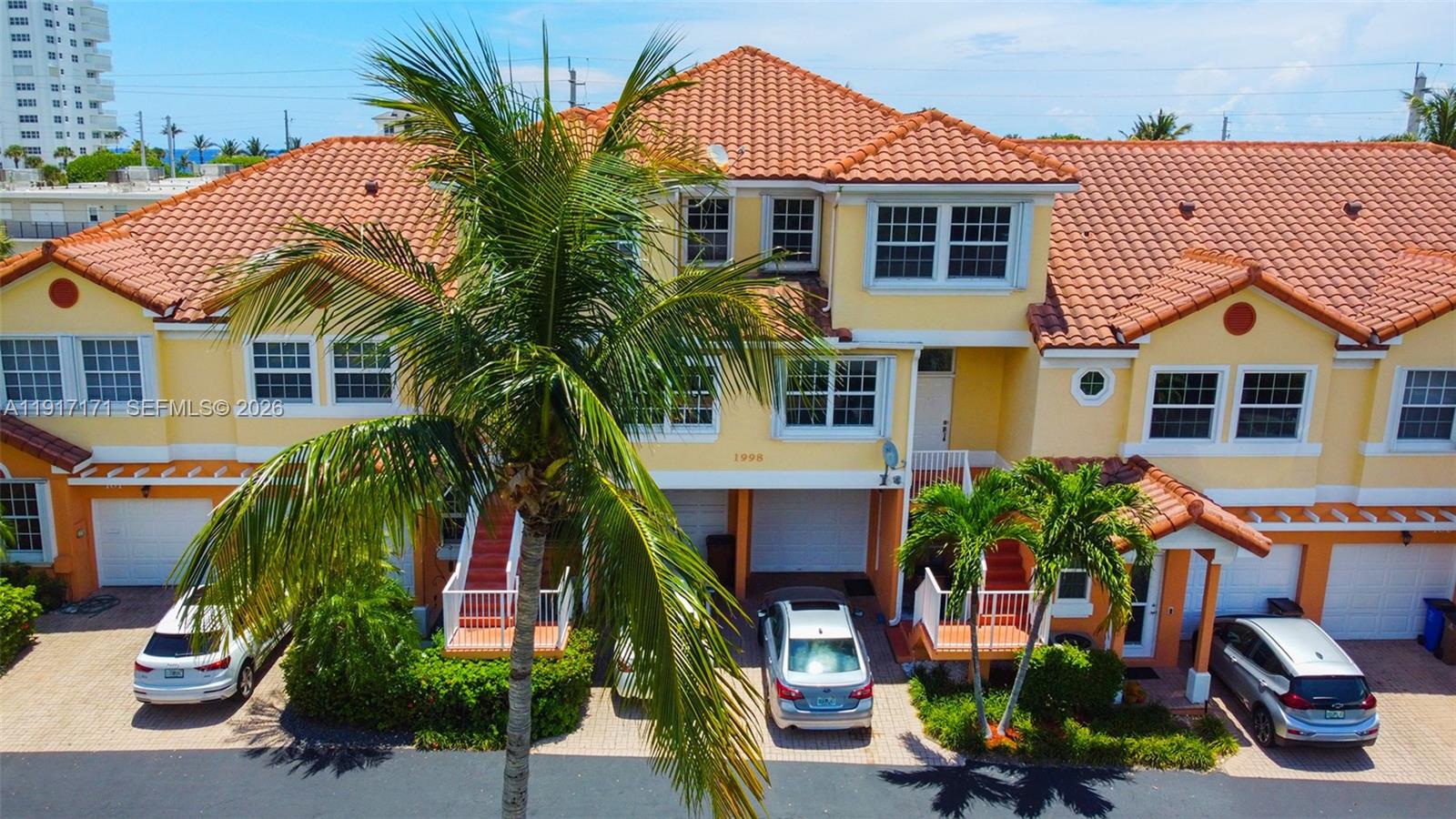 An exterior view of a multi-story yellow townhouse complex with a tiled roof and palm trees — 1998 NE 7th St # 104, Deerfield Beach FL 33441, DEERFIELD BEACH, FL