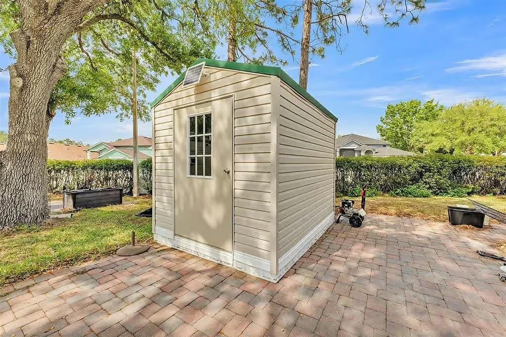 A beige vinyl storage shed with a green roof sits on a brick paver patio — 16329 CHERRY TREE COURT, CLERMONT, FL