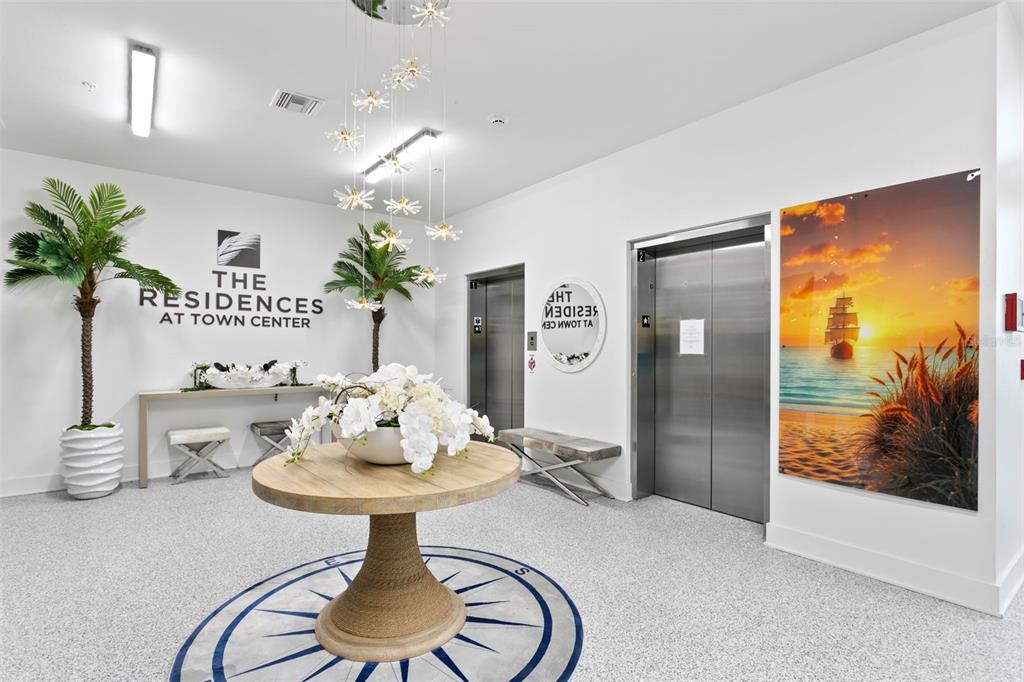 A modern apartment building lobby features a round table, two elevators, and a large ship painting — 400 150TH AVENUE UNIT 305, MADEIRA BEACH, FL