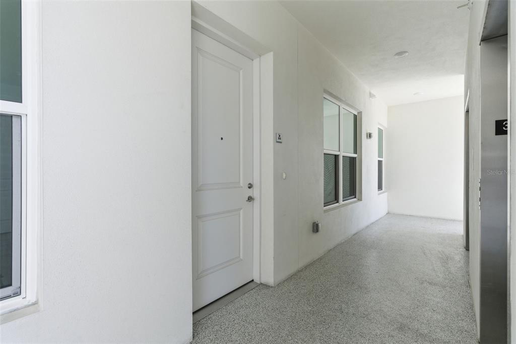 A white door leads into a unit from a bright, covered outdoor hallway with flooring — 400 150TH AVENUE UNIT 305, MADEIRA BEACH, FL