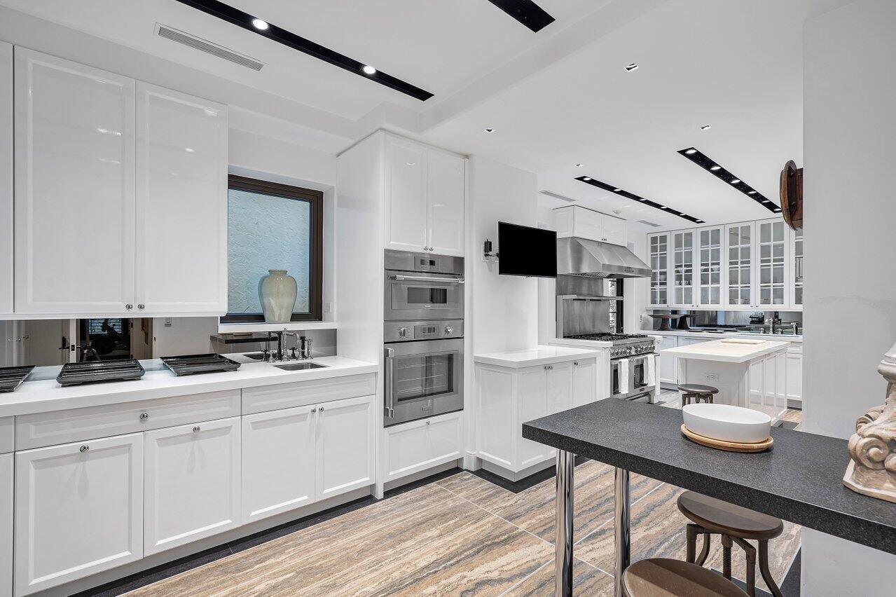 A modern white kitchen features stainless steel appliances, a center island, and wood-look tile flooring — 12 Lagomar Road, Palm Beach FL 33480, PALM BEACH, FL