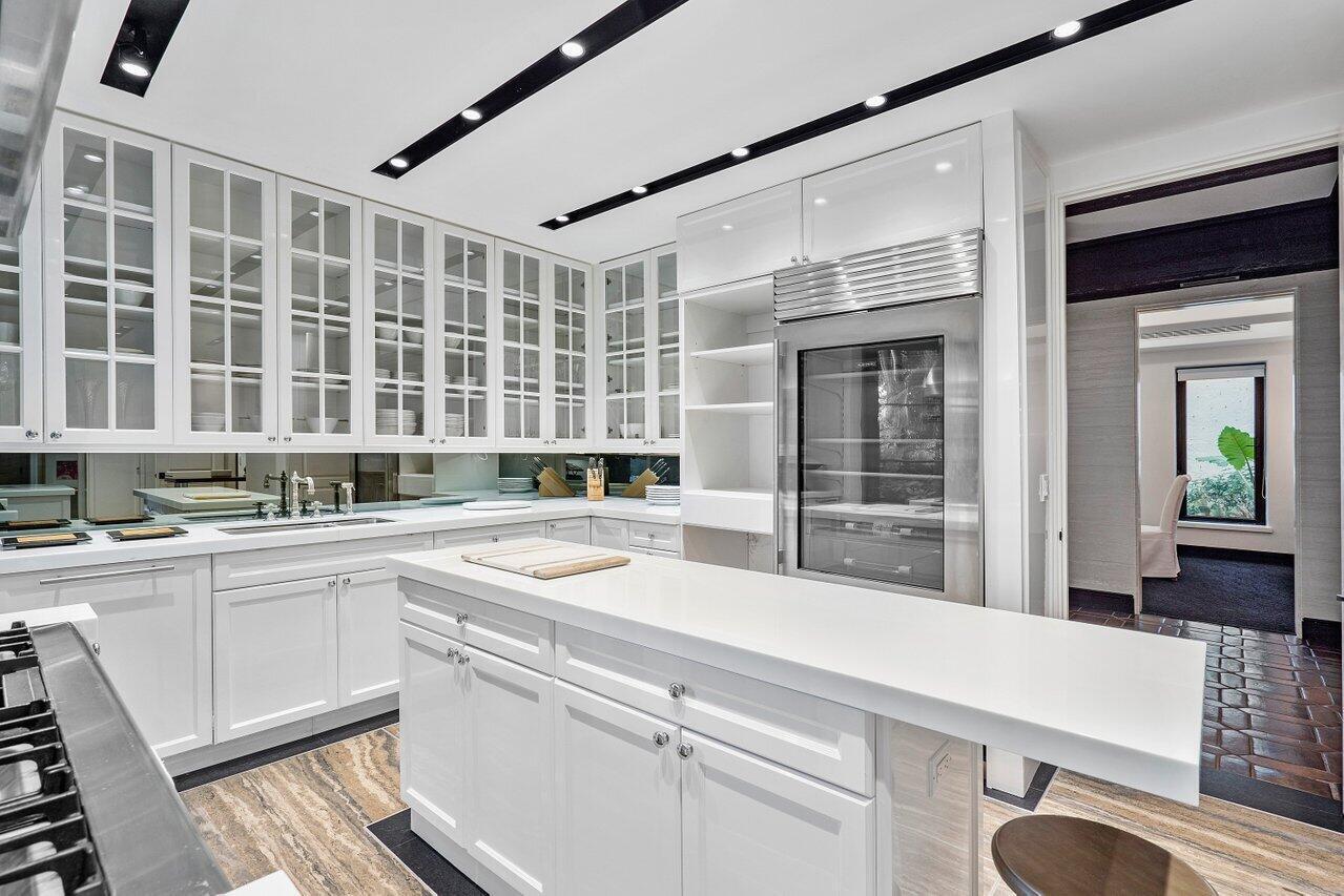 A white kitchen features a central island, glass-front cabinets, stainless steel appliances, and stone flooring — 12 Lagomar Road, Palm Beach FL 33480, PALM BEACH, FL
