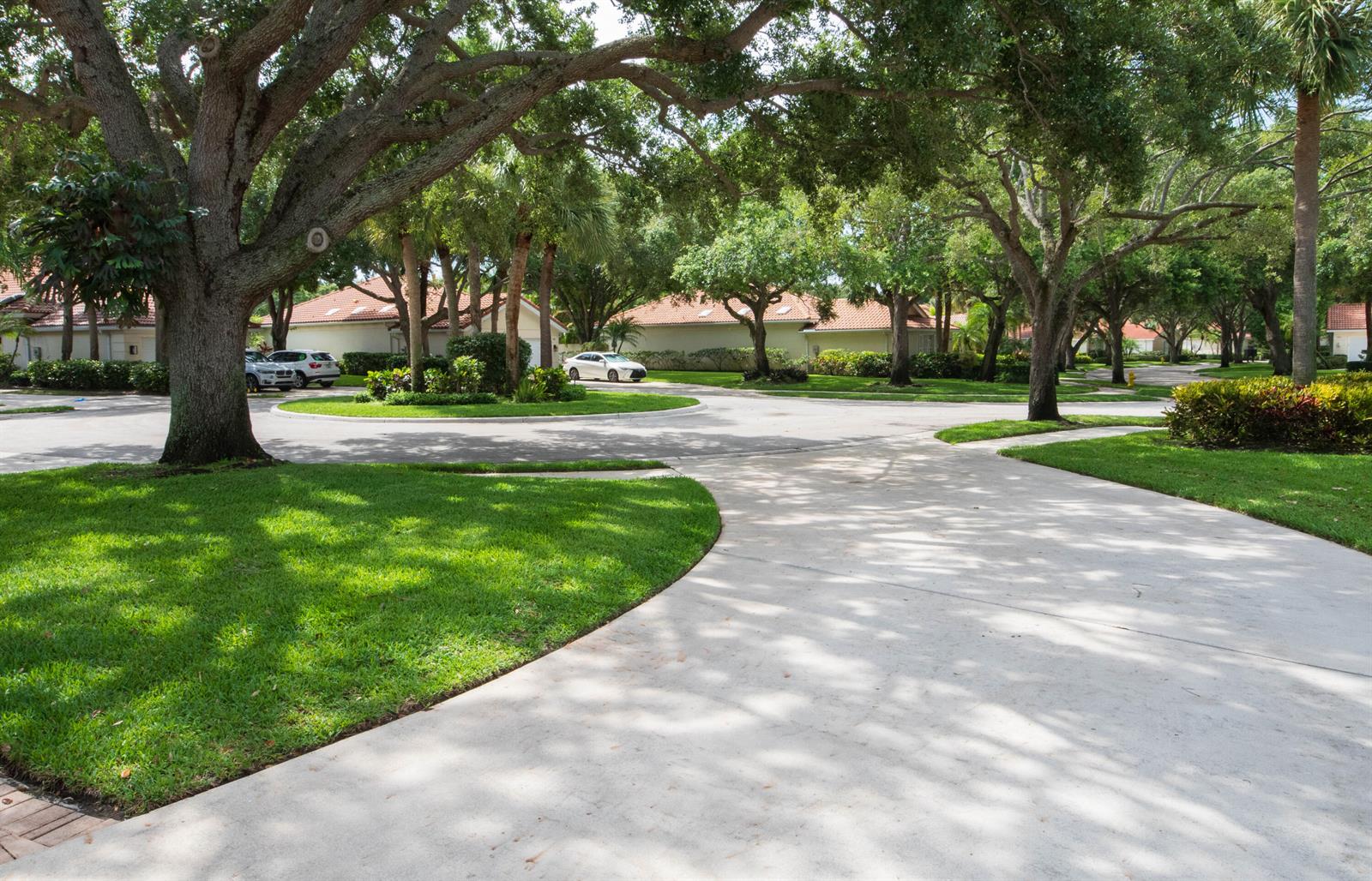 A paved driveway curves through a lush, tree-lined residential neighborhood with houses in the background — 146 Lost Bridge Drive, Palm Beach Gardens FL 33410, PALM BEACH GARDENS, FL