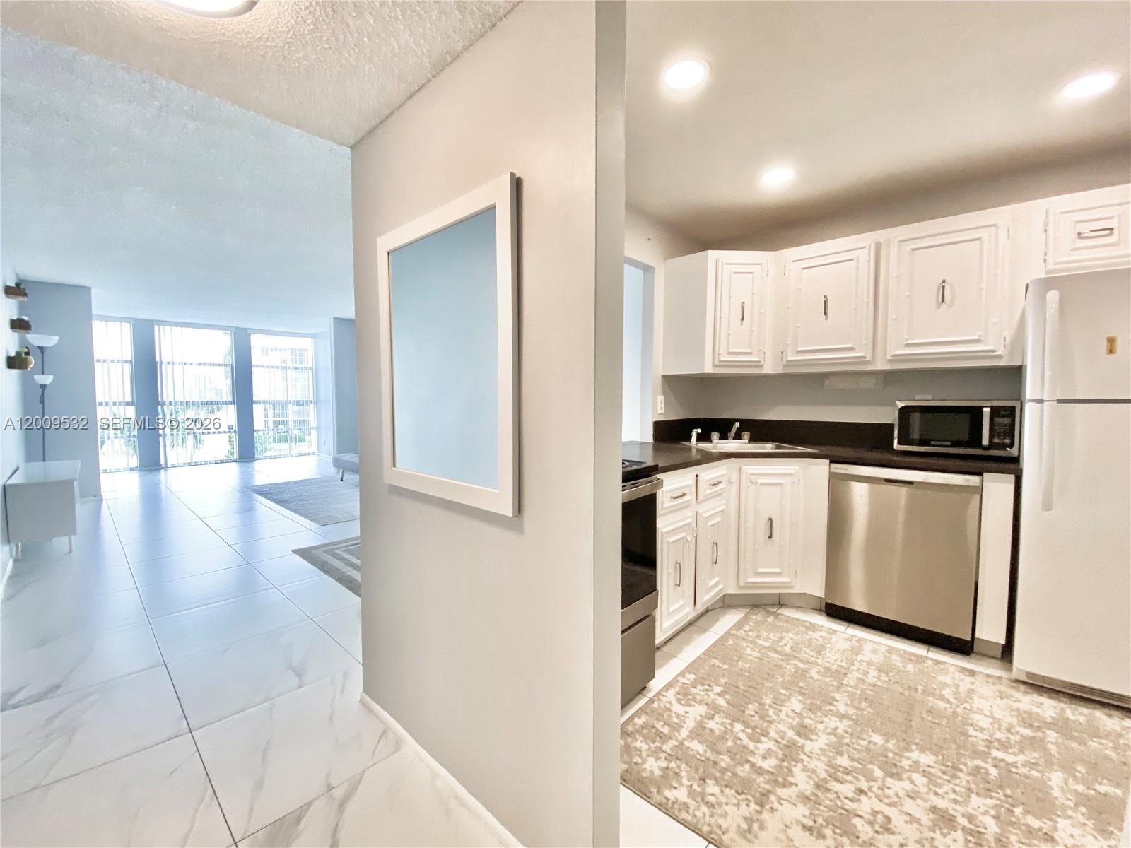 A kitchen with white cabinets and stainless steel appliances sits beside a tiled living area — 601 Three Islands Blvd # 314, Hallandale Beach FL 33009, HALLANDALE BEACH, FL