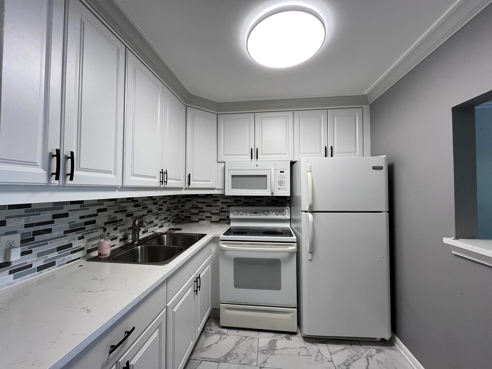 A kitchen features white cabinets, stainless steel sink, white appliances, and gray tiled backsplash — 49 Cambridge B, West Palm Beach FL 33417, WEST PALM BEACH, FL