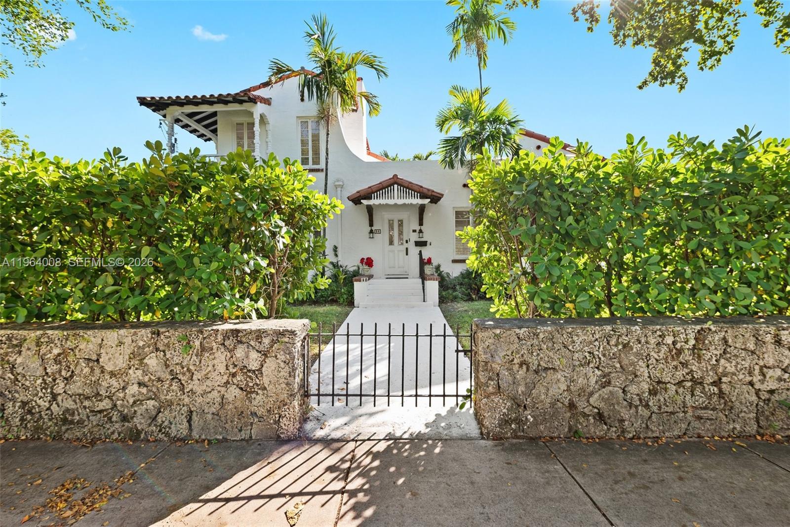 A white Mediterranean-style house features a stone wall, gated walkway, and lush green hedges — 36 Phoenetia Ave, Coral Gables FL 33134, CORAL GABLES, FL