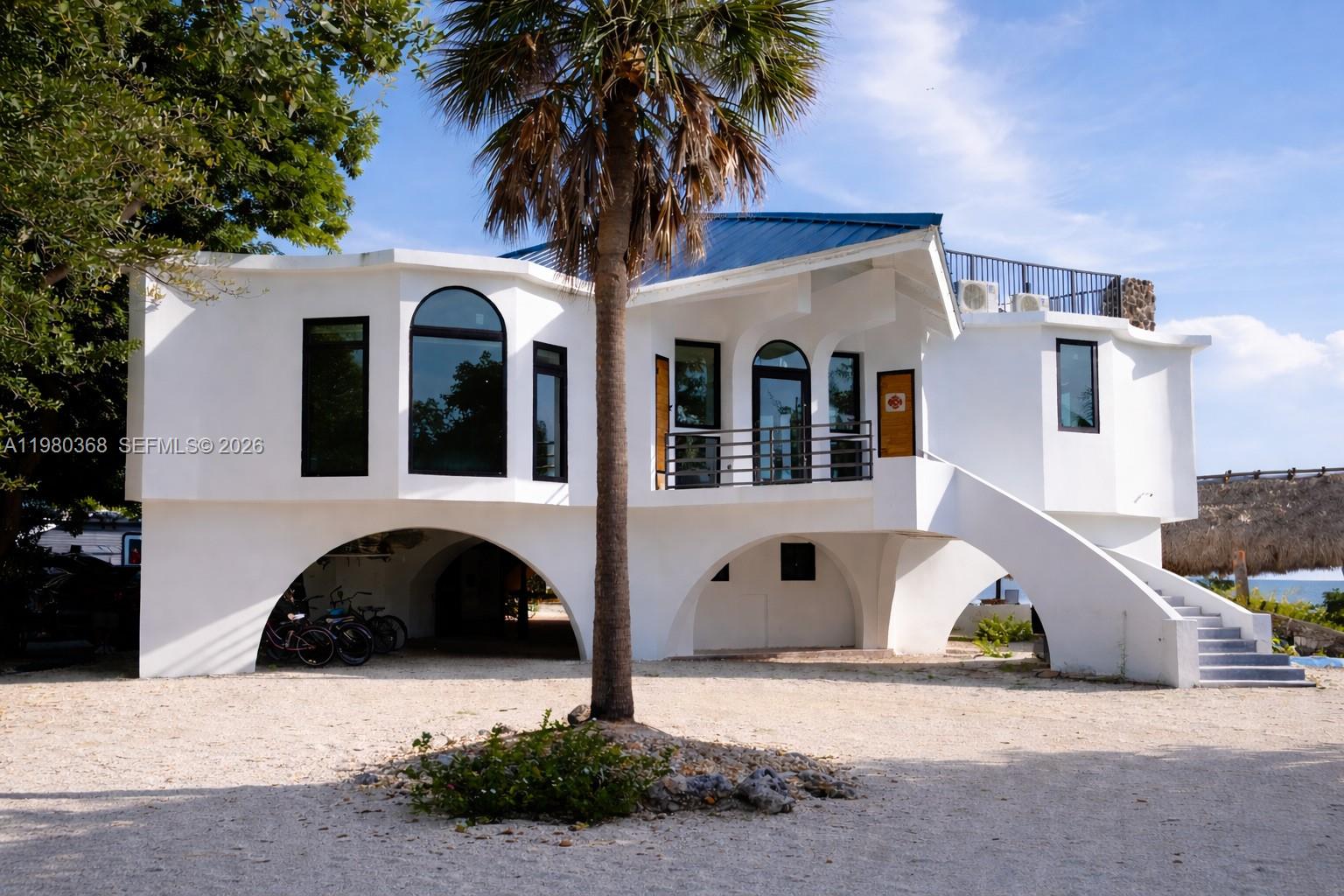 A white, elevated house with arched ground-level openings and an exterior staircase stands on gravel — 1557 Long Beach Dr, Big Pine FL 33043, BIG PINE, FL