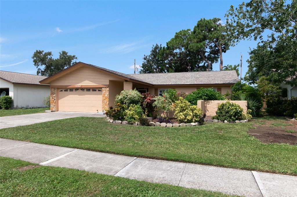 A single-story house with a tan exterior, a two-car garage, and a landscaped front yard — 1150 BROOK DRIVE W, DUNEDIN, FL