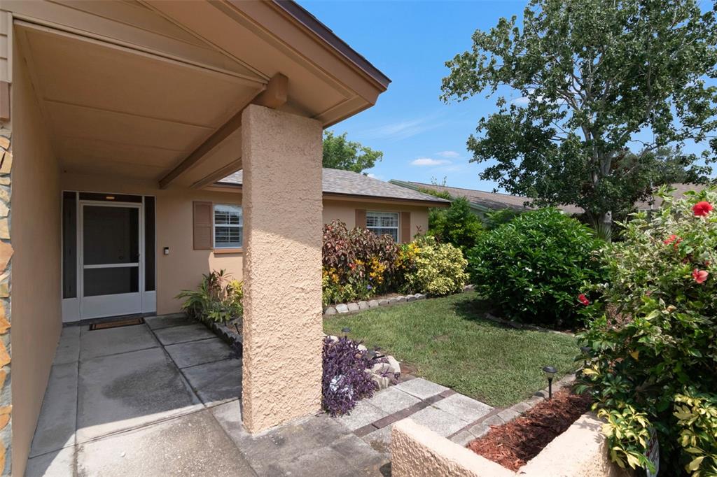 A beige house exterior features a covered entryway, a concrete walkway, and a landscaped yard — 1150 BROOK DRIVE W, DUNEDIN, FL