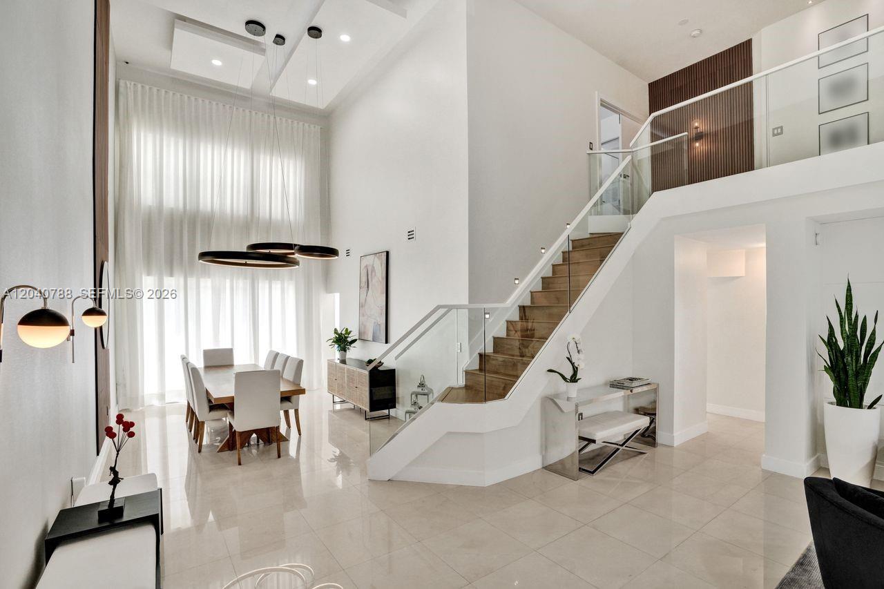 A modern, high-ceilinged dining area features a staircase with glass railings and tile flooring — 10762 NW 71st St, Doral FL 33178, DORAL, FL