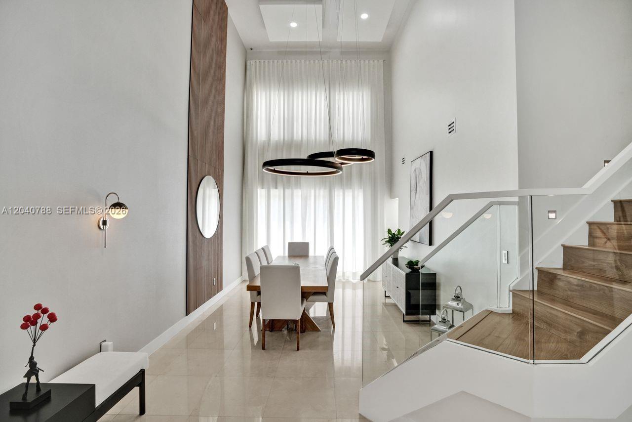 A modern dining room features a high ceiling, wood-slat accent wall, and glass-railed staircase — 10762 NW 71st St, Doral FL 33178, DORAL, FL