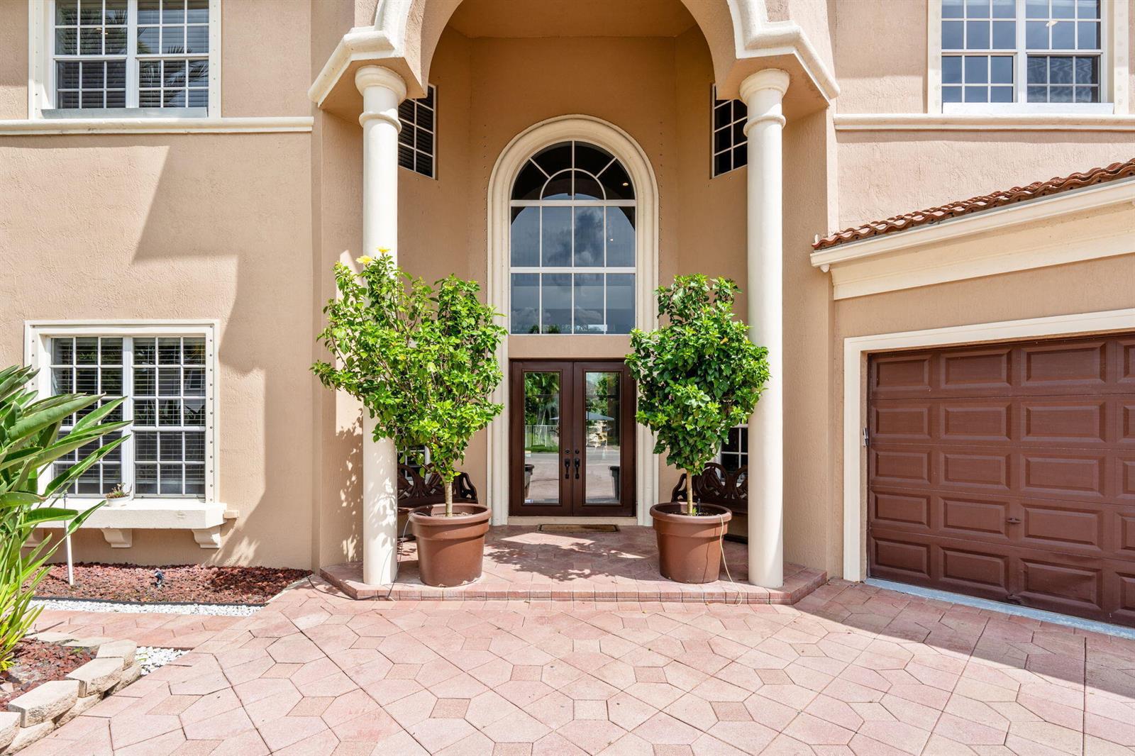 The exterior of a beige two-story house features a covered entryway with two potted trees — 12442 Equine Lane, Wellington FL 33414, WELLINGTON, FL