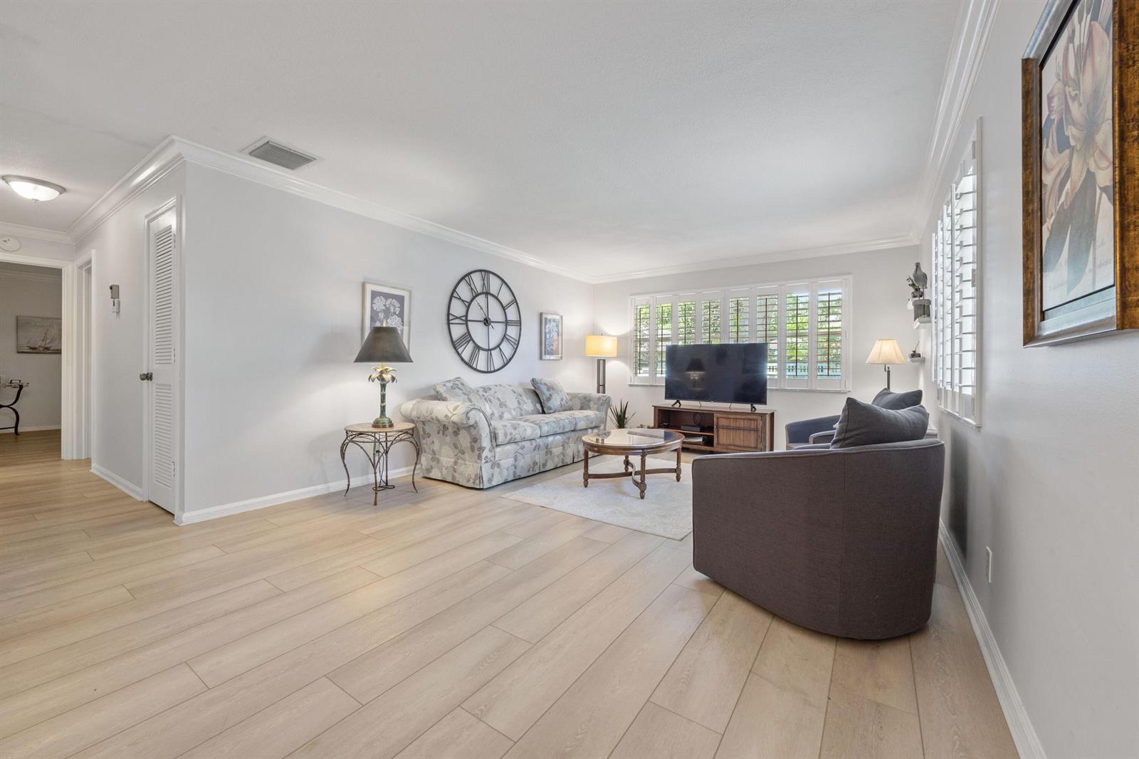 A living room features light wood flooring, a floral sofa, a television, and plantation shutters — 10116 46th Avenue S # 488, Boynton Beach FL 33436, BOYNTON BEACH, FL