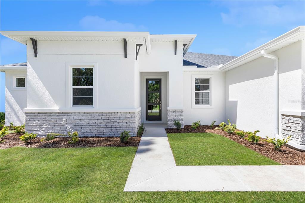 A modern white house exterior features stone veneer accents, a glass-paneled front door, and landscaping — 3518 NW 44TH PLACE, CAPE CORAL, FL