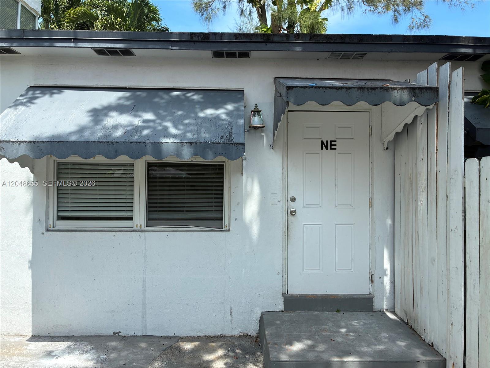 A white exterior wall features a window with a gray awning and a door labeled NE — 7929 Abbott Ave, Miami Beach FL 33141, MIAMI BEACH, FL