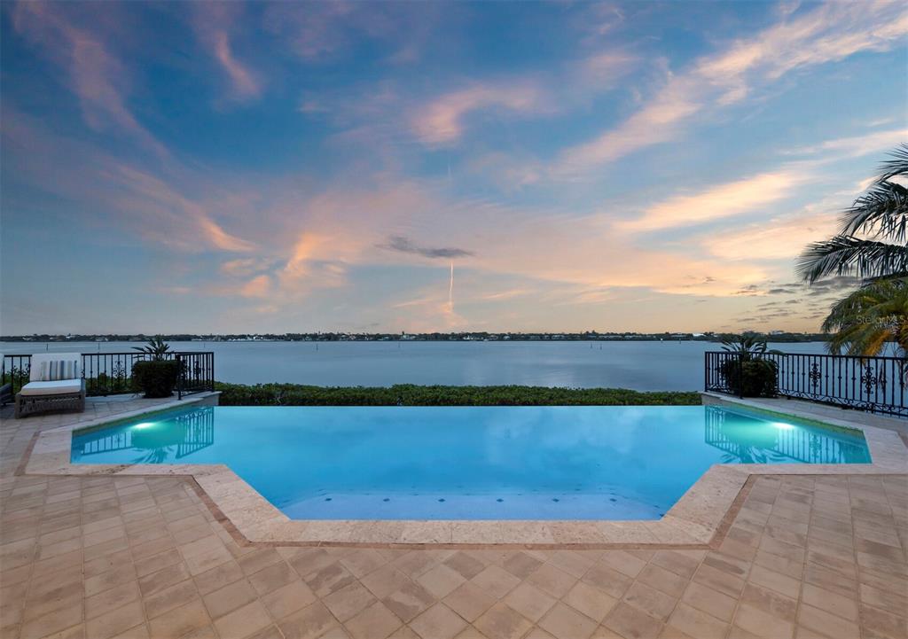 An infinity pool overlooks a large body of water at sunset from a stone patio — 1280 HIDDEN HARBOR WAY, SARASOTA, FL
