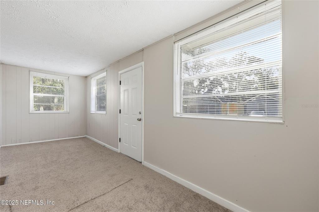An empty room features beige walls, carpeted flooring, three windows with blinds, and a door — 1217 W 29TH STREET, JACKSONVILLE, FL