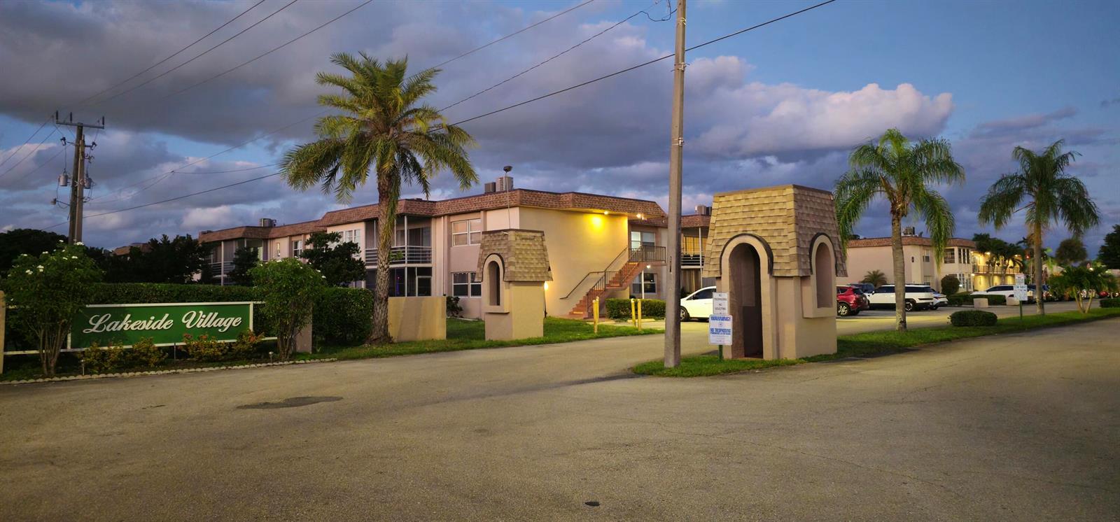 The exterior of the Lakeside Village apartment complex features a gated entrance and palm trees — 721 Lori Dr # 105, Palm Springs FL 33461, PALM SPRINGS, FL