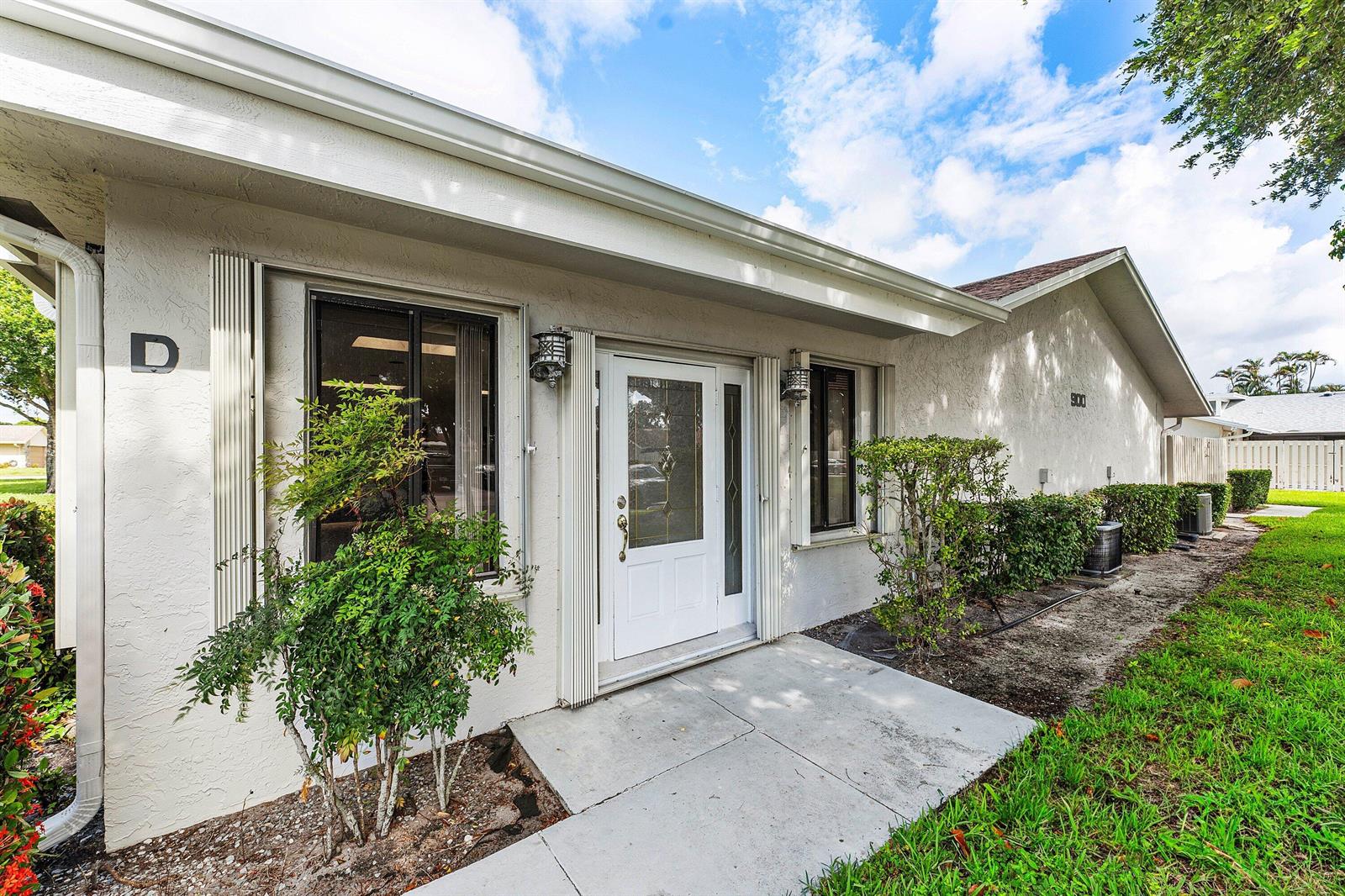 The exterior of a single-story building features a white front door, windows, and landscaping — 900 NW 30th Avenue # D, Delray Beach FL 33445, DELRAY BEACH, FL