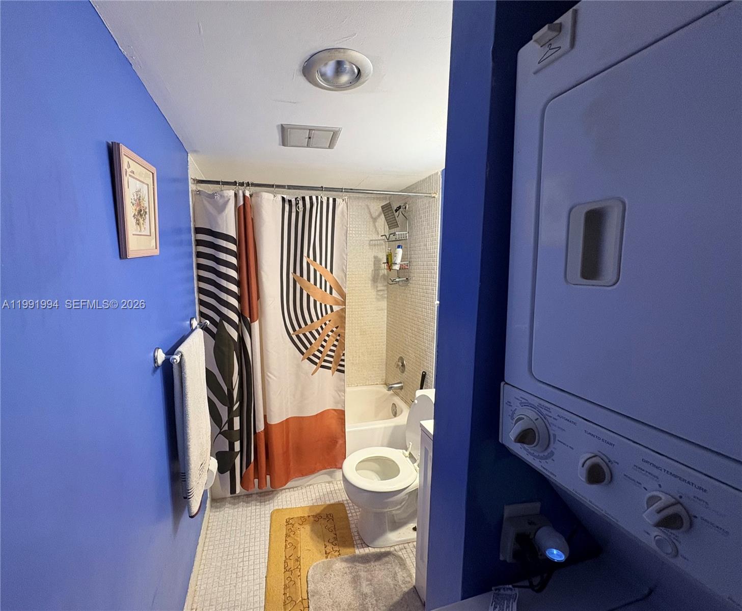 A bathroom with blue walls features a shower-tub combo, toilet, and a stacked laundry unit — 4152 Inverrary Dr # 303, Lauderhill FL 33319, LAUDERHILL, FL