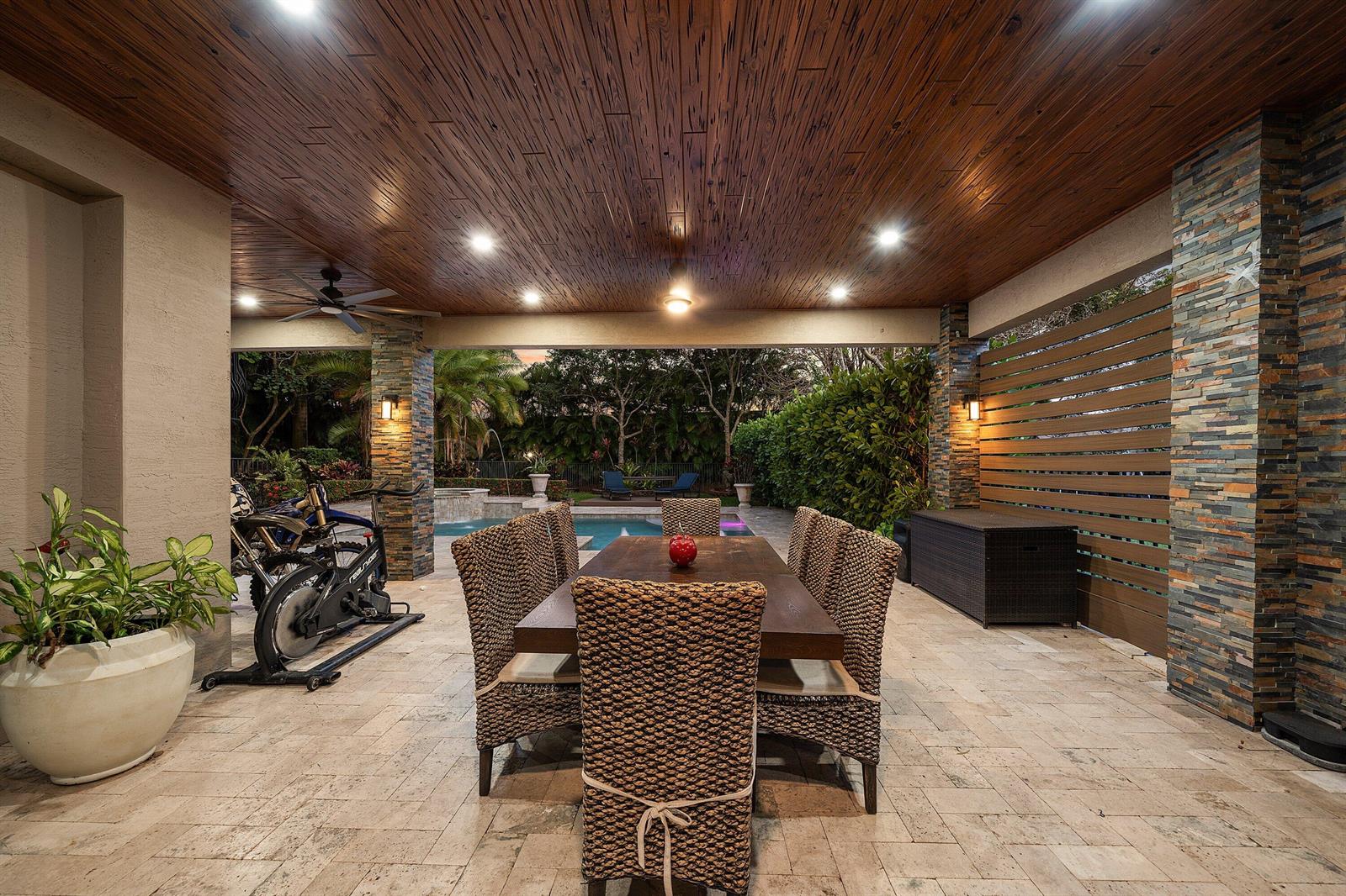 A covered outdoor patio features a dining set, exercise bike, stone pillars, and wood ceiling — 11565 Mantova Bay Circle, Boynton Beach FL 33473, BOYNTON BEACH, FL