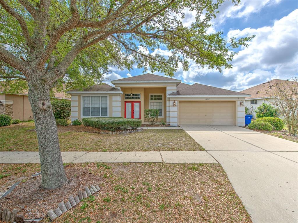 A single-story suburban house features a tan exterior, a red front door, and a driveway — 10907 AUSTRALIAN PINE DRIVE, RIVERVIEW, FL