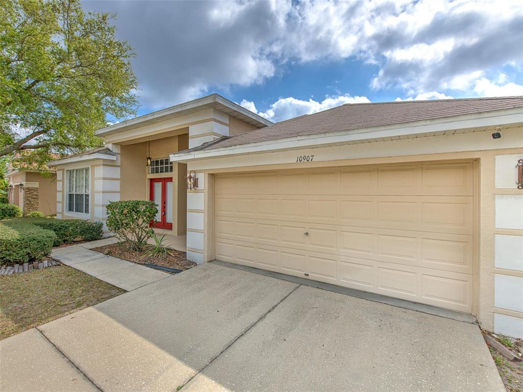 A beige single-story house features a two-car garage, a red front door, and landscaping — 10907 AUSTRALIAN PINE DRIVE, RIVERVIEW, FL