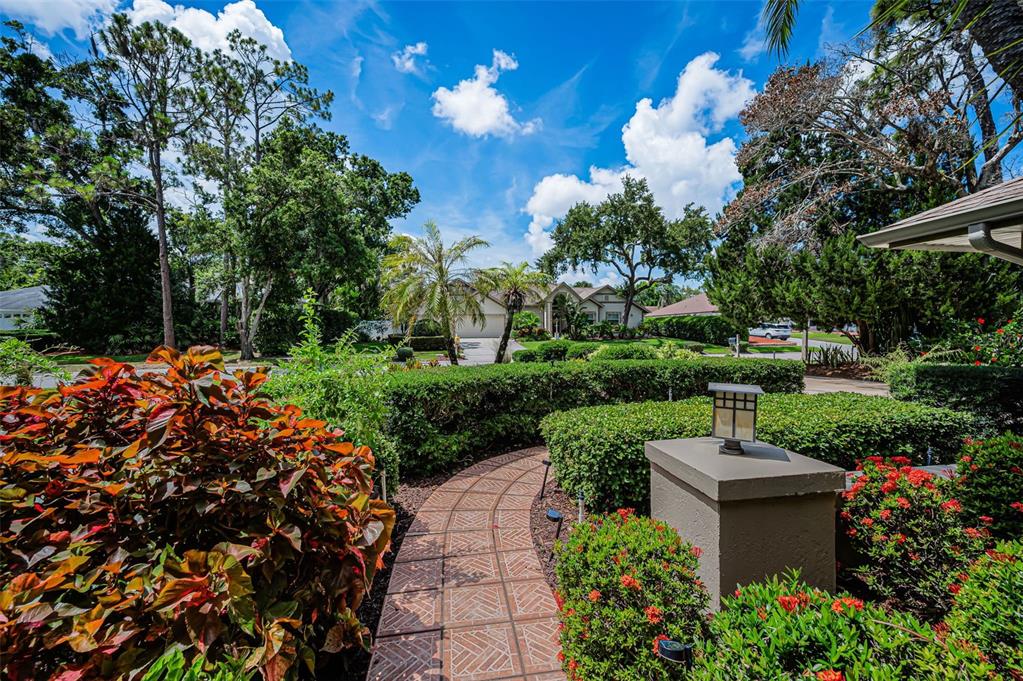 A tiled walkway leads through a landscaped yard toward a house under a blue sky — 1680 SPARKLING COURT, DUNEDIN, FL