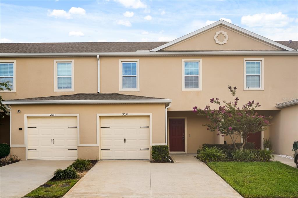 A two-story townhouse exterior with two garage doors, beige stucco walls, and a red door — 9031 WILDFLOWER LANE, KISSIMMEE, FL