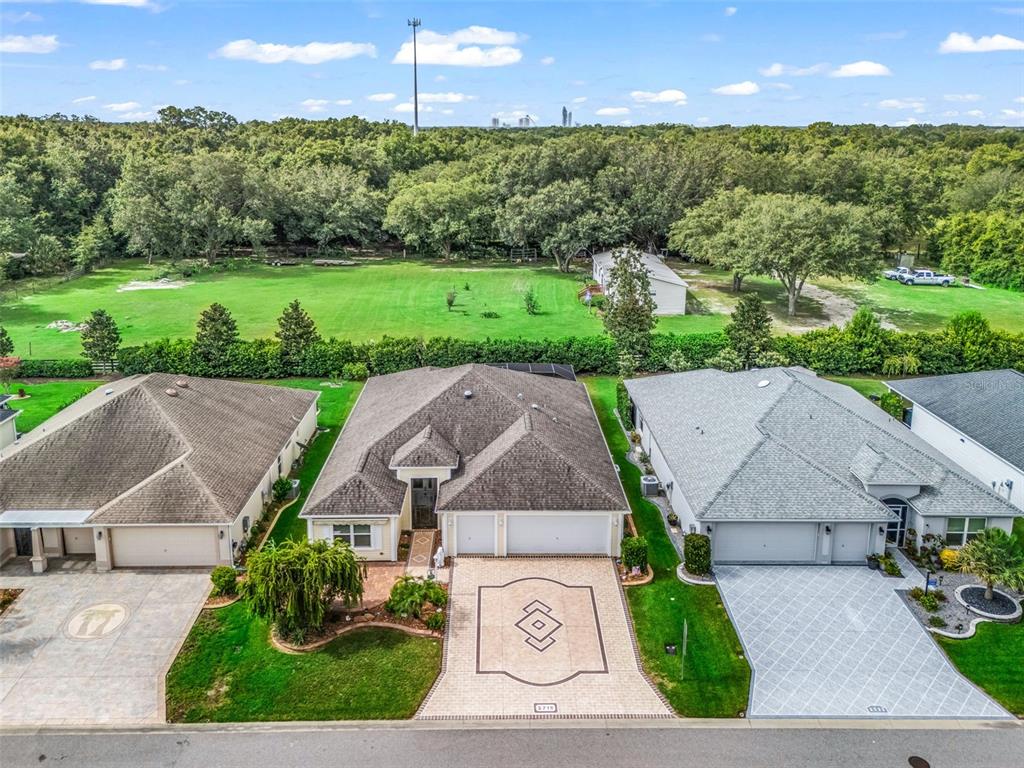 An aerial view shows three suburban houses with paved driveways and a large grassy backyard — 2716 LYALL LOOP, THE VILLAGES, FL