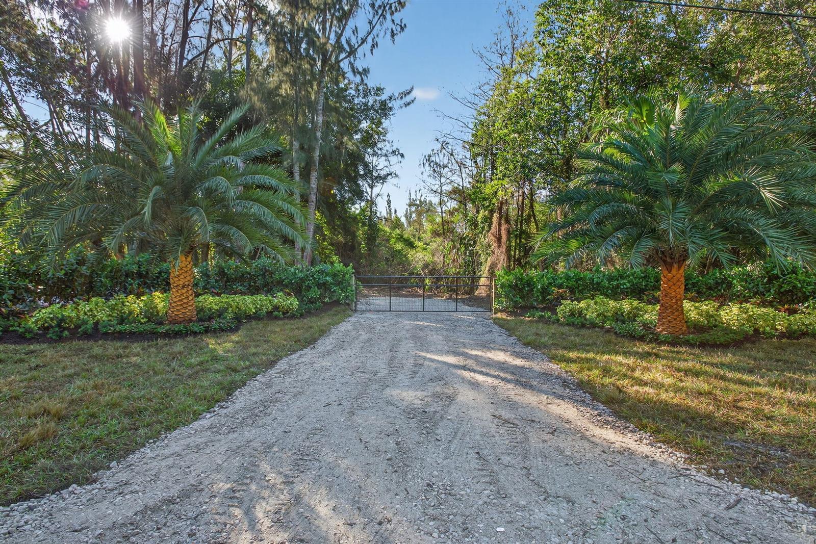 A gravel driveway leads to a metal gate flanked by two palm trees and landscaping — 9620 171st Street N, Jupiter FL 33478, JUPITER, FL