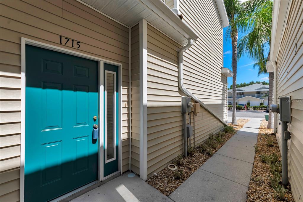 A teal front door with a side window is set into a beige-sided building exterior — 1715 HAMMOCKS AVENUE UNIT 1715, LUTZ, FL