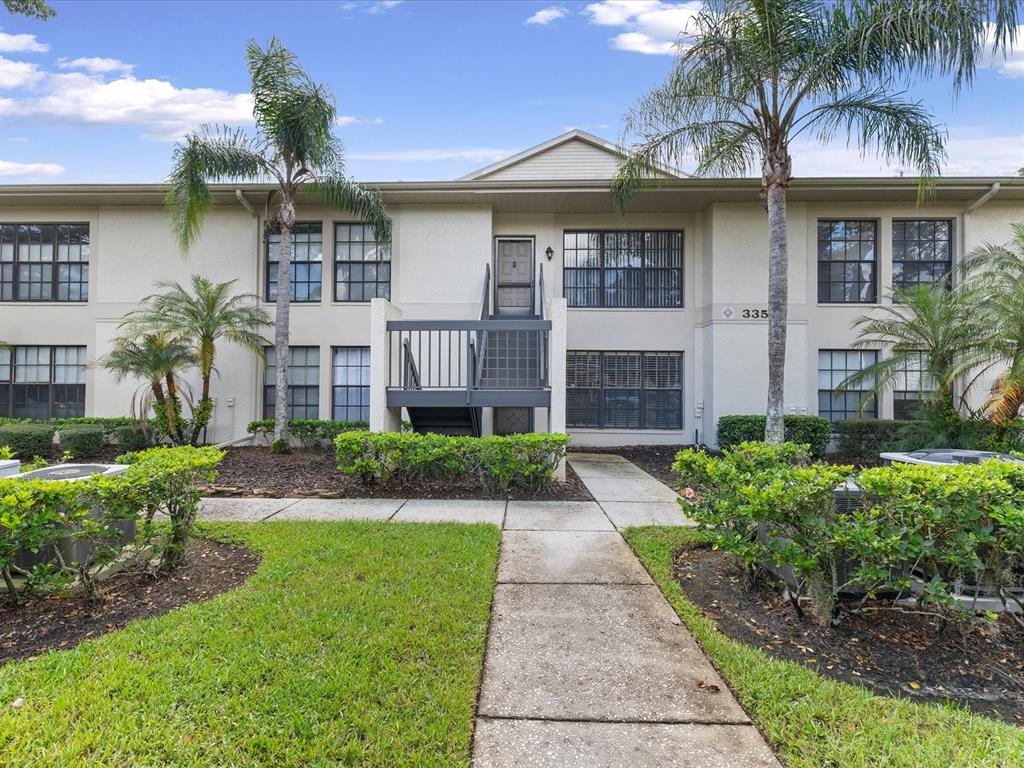 A two-story apartment building exterior features a central staircase, beige siding, and palm trees — 3358 MERMOOR DRIVE UNIT 102, PALM HARBOR, FL