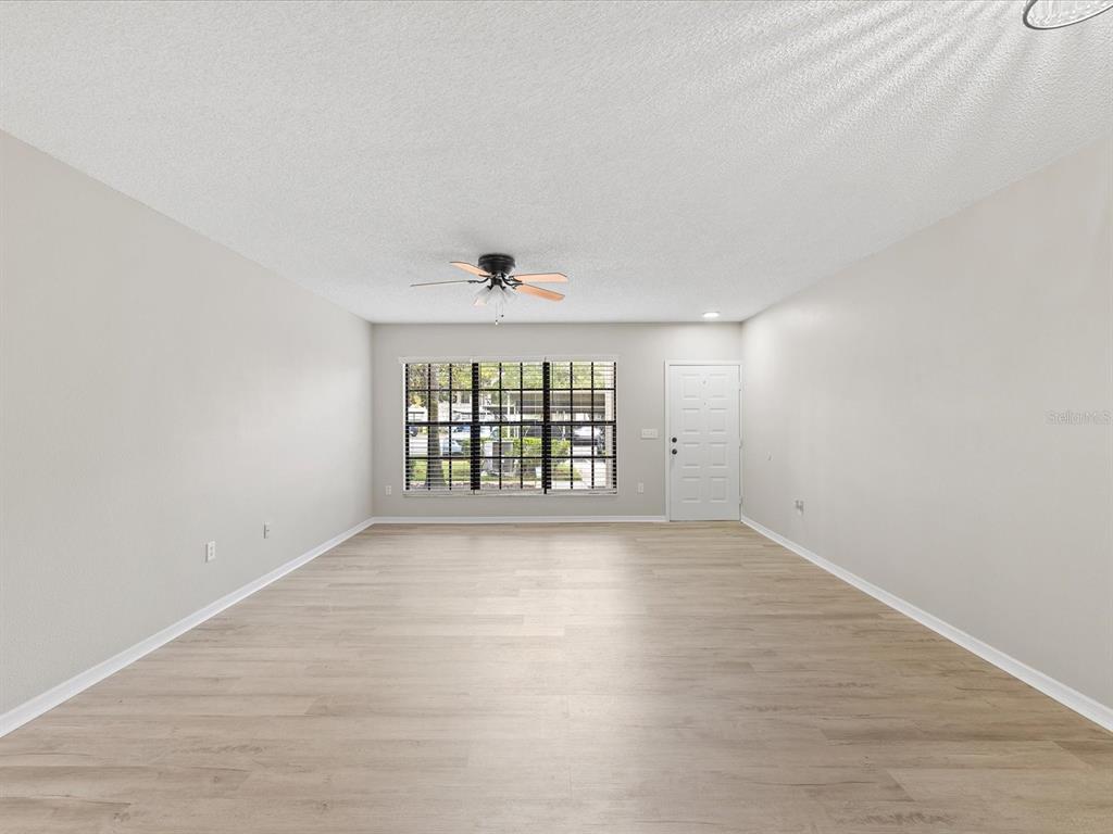 An empty living room features light-colored wood-look flooring, white walls, a ceiling fan, and windows — 3358 MERMOOR DRIVE UNIT 102, PALM HARBOR, FL